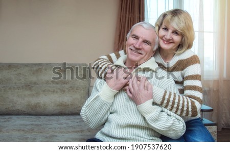 A married couple of 50-60 years old are hugging while sitting on the sofa at home. Looking into the camera. The concept of a long happy marriage. Place for text. Royalty-Free Stock Photo #1907537620