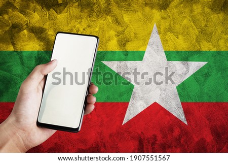 Hand holding smartphone on Myanmar Flag background.