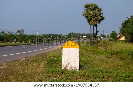Captured Real Photo of Plain Yellow and White Milestone with National Highway Road and green plants Background which can be used for writing Kilometre numbers on it found in Tamilnadu South India Asia Royalty-Free Stock Photo #1907554357