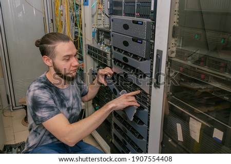 A young man works in a server room. A technician maintains computer equipment in a data center. An engineer works near the data server racks.