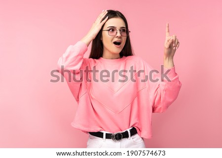 Photo of attractive young woman in glasses recalls something, got idea. Wears casual pink t-shirt white pants isolated pink color background