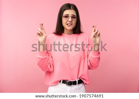 Photo of attractive young woman in glasses cross fingers, insecure and worried. Wears casual pink t-shirt white pants isolated pink color background