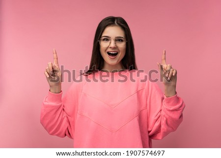Photo of attractive young woman in glasses points up and surprised. Wears casual pink t-shirt isolated pink color background
