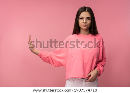 Photo of attractive young woman in glasses, serious face and shows refuse. Wears casual pink t-shirt white pants isolated pink color background