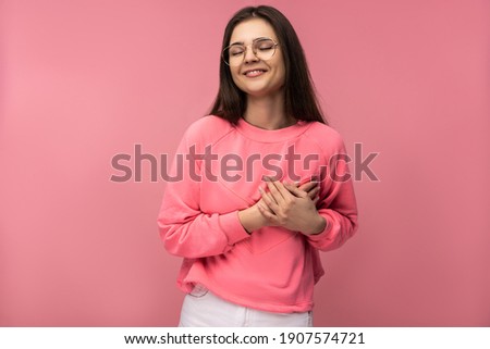 Photo of attractive young woman in glasses holds heart. Wears casual pink t-shirt white pants isolated pink color background