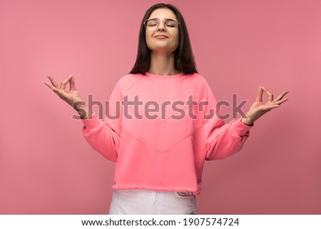 Photo of attractive young woman in glasses, meditation and relax, wears casual pink t-shirt white pants isolated pink color background