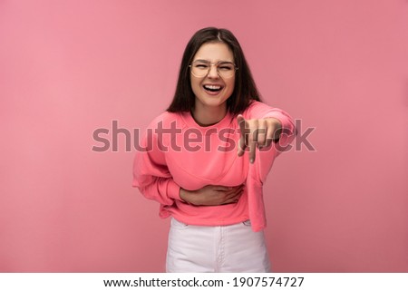 Photo of attractive young woman in glasses laughs pointing at camera, wears casual pink t-shirt white pants isolated pink color background