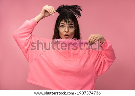 Photo of attractive young woman in glasses holds feathers with surprised, wears casual pink t-shirt white pants isolated pink color background