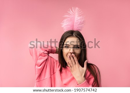 Photo of attractive young woman in glasses holds feathers back head, covers mouth and surprised. Wears casual pink t-shirt white pants isolated pink color background