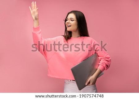 Photo of attractive lady in glasses says hi and hold laptop, study online. Wears casual pink t-shirt white pants isolated pink color background