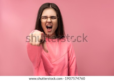 Photo of attractive young woman in glasses yelling and pointing at camera. Wears casual pink t-shirt white pants isolated pink color background