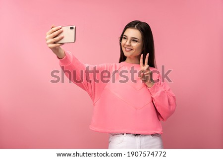 Photo of attractive young woman in glasses takes selfie photo on smartphone, wears casual pink t-shirt white pants isolated pink color background