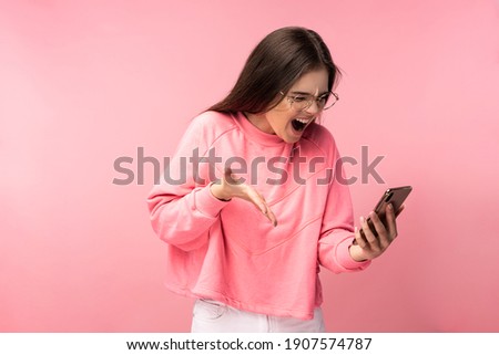 Photo of attractive lady in glasses holds telephone and screams, disappointed reads comments on blog. Wears casual pink t-shirt white pants isolated pink color background.