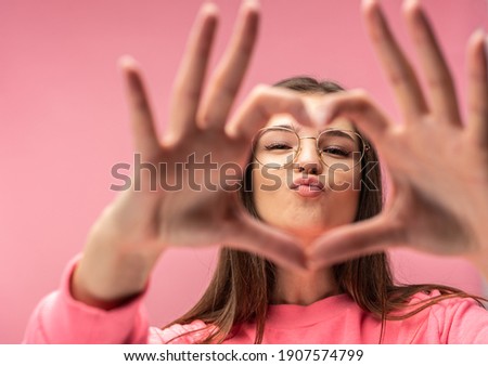 Photo of attractive young woman in glasses shows heart with fingers, blow kiss. Wears casual pink t-shirt white pants isolated pink color background