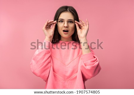 Photo of attractive young woman in glasses looks unexpectedly and surprised, wears casual pink t-shirt white pants isolated pink color background