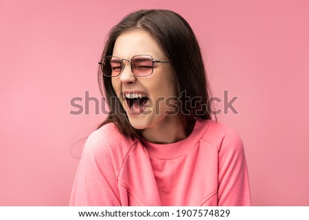 Photo of attractive young woman in glasses sings, screams and laughs. Wears casual pink t-shirt white pants isolated pink color background