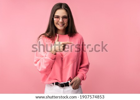 Photo of attractive young woman in glasses points at right and smile, wears casual pink t-shirt white pants isolated pink color background