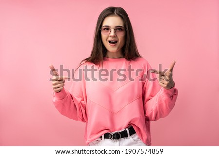 Photo of attractive young woman in glasses points at something and surprised, amazed. Wears casual pink t-shirt white pants isolated pink color background