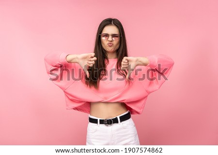 Photo of attractive young woman in glasses dislike and disagree with something. Wears casual pink t-shirt white pants isolated pink color background.
