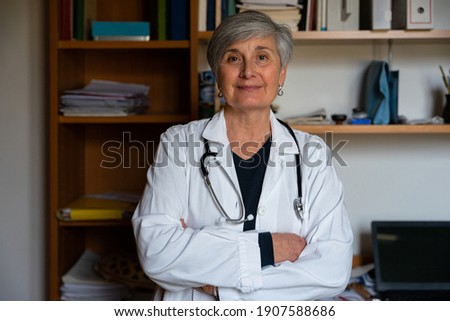 Portrait of a female family doctor in her office with her arms crossed - 60s woman with white uniform in her work space