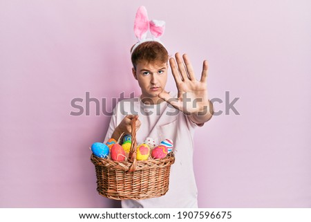 Young caucasian man wearing cute easter bunny ears holding wicker basket with colored eggs with open hand doing stop sign with serious and confident expression, defense gesture 