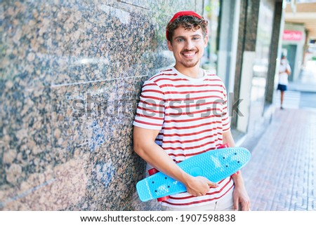 Young caucasian skater man smiling happy holding skate leaning on the wall city.