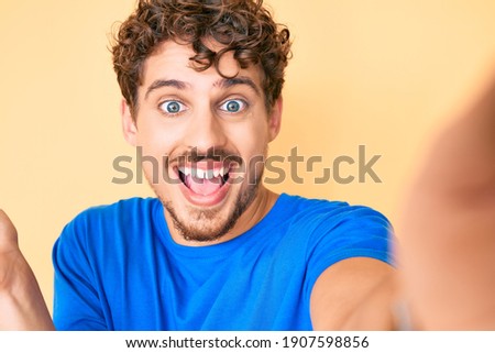 Young caucasian man with curly hair wearing casual clothes taking a selfie celebrating victory with happy smile and winner expression with raised hands 