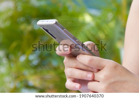 South east asian Female hands are holding mobile smart phone with blurred green color palm tree background with blank empty copy space for tex or background usage 