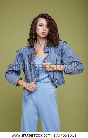 High fashion photo of a beautiful elegant young woman in a pretty denim jeans jacket, blue trousers, white boots, posing over green background. Studio Shot. Portrait