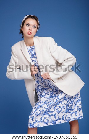High fashion photo of a beautiful elegant young woman in a pretty white jacket, patterned dress, boots, head scarf posing over blue background. Studio Shot. Portrait