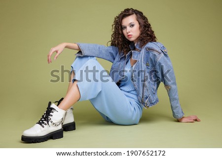 High fashion photo of a beautiful elegant young woman in a pretty denim jeans jacket, blue trousers, white boots, posing over green background. Studio Shot. Portrait. Model is sitting