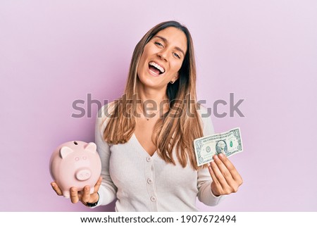 Brunette young woman holding one dollar banknote and piggy bank smiling and laughing hard out loud because funny crazy joke. 
