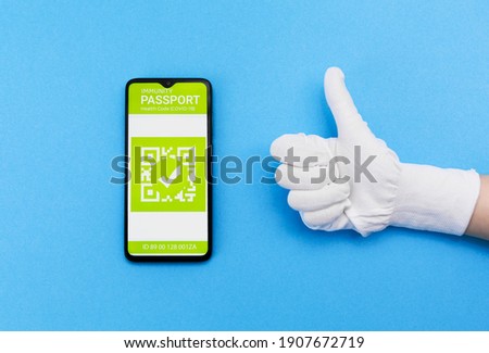 Concept of vaccination and digital passport. Hand in gloves shows thumb up and electronic Immunity passport with a COVID-19 vaccination stamp on a smartphone screen on the blue background. Flat lay. Royalty-Free Stock Photo #1907672719