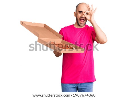 Young handsome man holding delivery pizza cardboard box smiling happy doing ok sign with hand on eye looking through fingers 