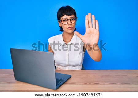 Beautiful brunettte woman working using computer laptop with open hand doing stop sign with serious and confident expression, defense gesture 