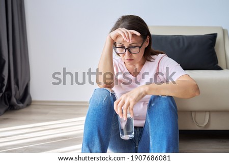 Sad middle aged woman sitting at home on the floor with glass of water. Health problems of older women, mental health, migraine pain, menopause symptom, depression period after coronavirus covid-19 Royalty-Free Stock Photo #1907686081