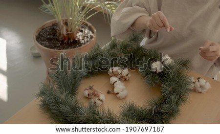European female florist in a beige sweatshirt sitting at the wooden table preparing cotton flower to put on the Chrstmas wreath. Warm light atmosphere. Hands shot down view photo