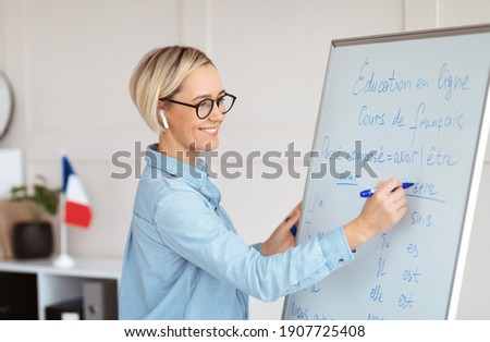 Friendly female teacher writing grammar rules on blackboard during online French lesson. Positive young tutor teaching foreign language on web. Remote education and tutoring concept Royalty-Free Stock Photo #1907725408