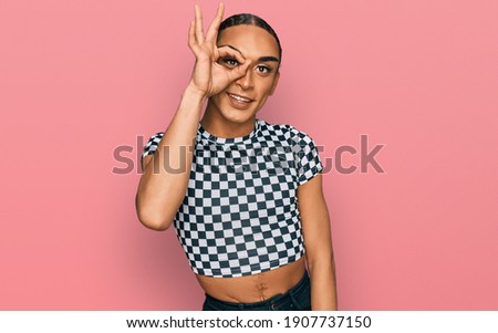 Hispanic transgender man wearing make up and long hair wearing modern clothes doing ok gesture with hand smiling, eye looking through fingers with happy face. 