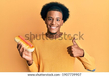 African american man with afro hair eating hotdog smiling happy and positive, thumb up doing excellent and approval sign 