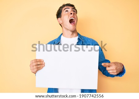 Young handsome man holding blank empty banner angry and mad screaming frustrated and furious, shouting with anger looking up. 