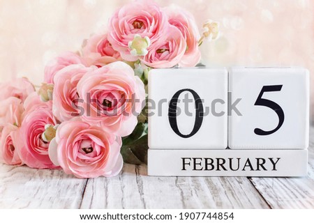 White wood calendar blocks with the date February 5th and pink ranunculus flowers over a wooden table. Selective focus with blurred background. 
