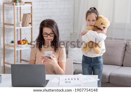 Lonely teen girl hugging teddy bear while her busy mom working online from home, not paying attention to child. Depressed kid feeling neglected, missing her parent. Covid-19 quarantine family problems Royalty-Free Stock Photo #1907754148