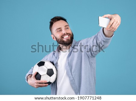 Positive millennial guy with soccer ball taking selfie on smartphone over blue studio background. Joyful football fan making photo of himself before championship, rooting for his favorite team