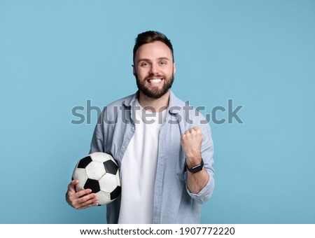 Joyful young guy holding soccer ball, making YES gesture, happy over win of his favorite team on blue studio background. Portrait of football rooter during world championship. Sports and leisure