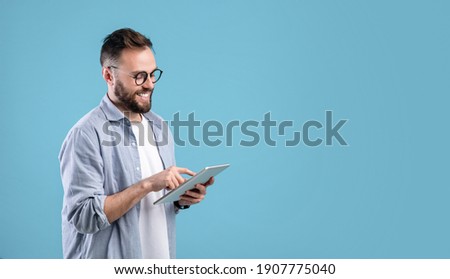 Smart millennial guy in glasses using tablet computer over blue studio background, banner design with copy space. Cheerful young man working, studying or communicating online