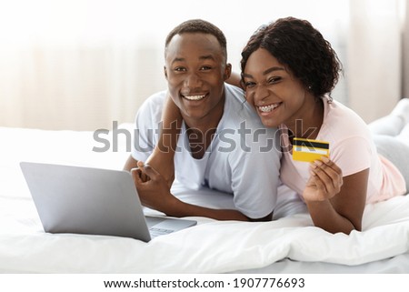 Young black couple laying on bed with laptop, shopping online, recommending credit card, closeup, copy space. Smiling african american man and woman in pajamas making online order, using computer