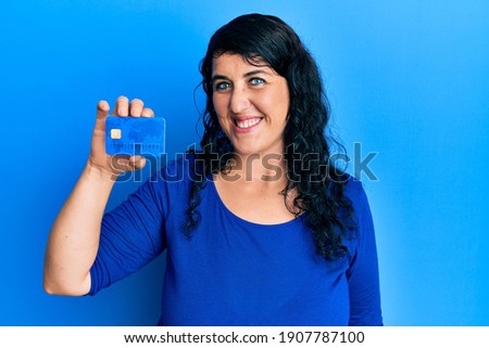 Plus size brunette woman holding credit card looking positive and happy standing and smiling with a confident smile showing teeth 