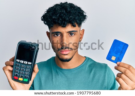 Young arab handsome man holding dataphone and credit card relaxed with serious expression on face. simple and natural looking at the camera. 