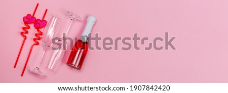Two champagne glasses with red plastic drinking straws with hearts and red champagne bottle on a pink background.